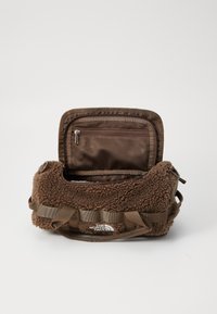 Brown fur-textured bag with a zippered mesh pocket inside, reinforced straps, and a front logo. Compact cylindrical shape.