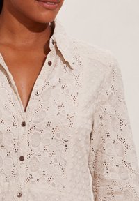 Camicia di cotone color crema con intricata ricamo in pizzo, dotata di chiusura a bottoni frontale, colletto a punte e motivi floreali testurizzati.