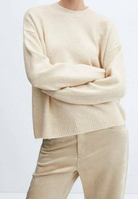 Pull beige en maille avec un col rond, des épaules tombantes et des poignets côtelés. Porté par-dessus un pantalon en velours côtelé beige clair. Texture douce.