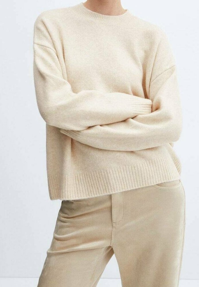 Pull beige en maille avec un col rond, des épaules tombantes et des poignets côtelés. Porté par-dessus un pantalon en velours côtelé beige clair. Texture douce.
