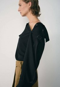 Schwarze Bluse mit weitem Kragen, langen Ärmeln und Knopfakzenten. Der Stoff wirkt glatt und fällt locker über einen beigen, hoch taillierten Rock.