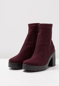 Burgunderfarbene Ankle Boots aus strukturiertem Material mit einem hohen Blockabsatz und einer dicken Plateausohle. Nahtdetails an den Seiten.