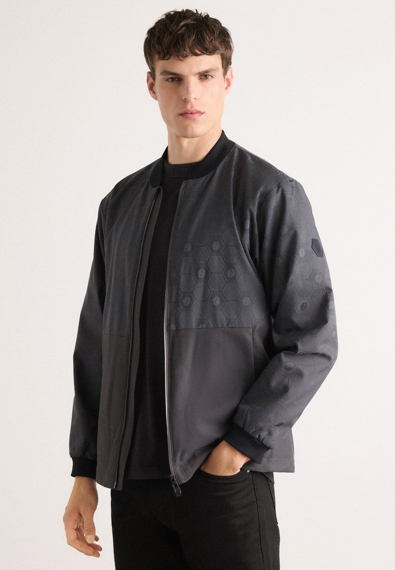 Bomber jacket in black with a hexagonal pattern on the upper left, smooth texture, zip closure, ribbed cuffs and collar.