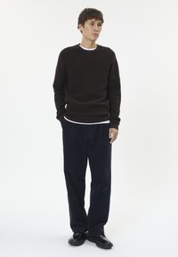 Maglione marrone scuro con texture a coste, indossato sopra una camicia bianca. I pantaloni in corduroy blu navy e le scarpe in pelle nere completano l'outfit.