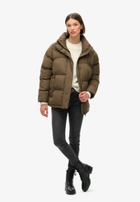 Puffjakke i olivengrøn, med høj krave, trykknapper og quilted tekstur. Sammen med en cremet sweater, mørke jeans og sorte støvler.