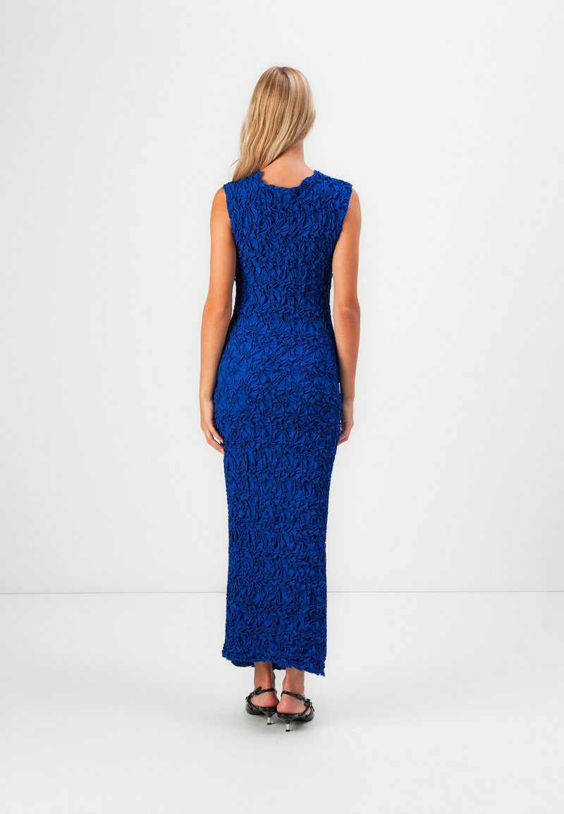 Kobieta z długimi blond włosami, ubrana w bezrękawnikową, teksturowaną, królewskoniebieską sukienkę maxi oraz czarne sandały na niskim obcasie, stoi przy białej ścianie.