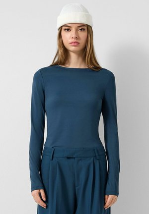 Jonge vrouw met een witte gebreide muts en een blauwe top met lange mouwen, gecombineerd met bijpassende blauwe hoge taille broek, staande tegen een effen muur.