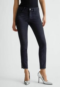 Jeans ajustados de mezclilla negro de cintura alta, con aberturas laterales en los tobillos y detalles de herrajes plateados. Combinados con zapatos de tacón alto metálicos.