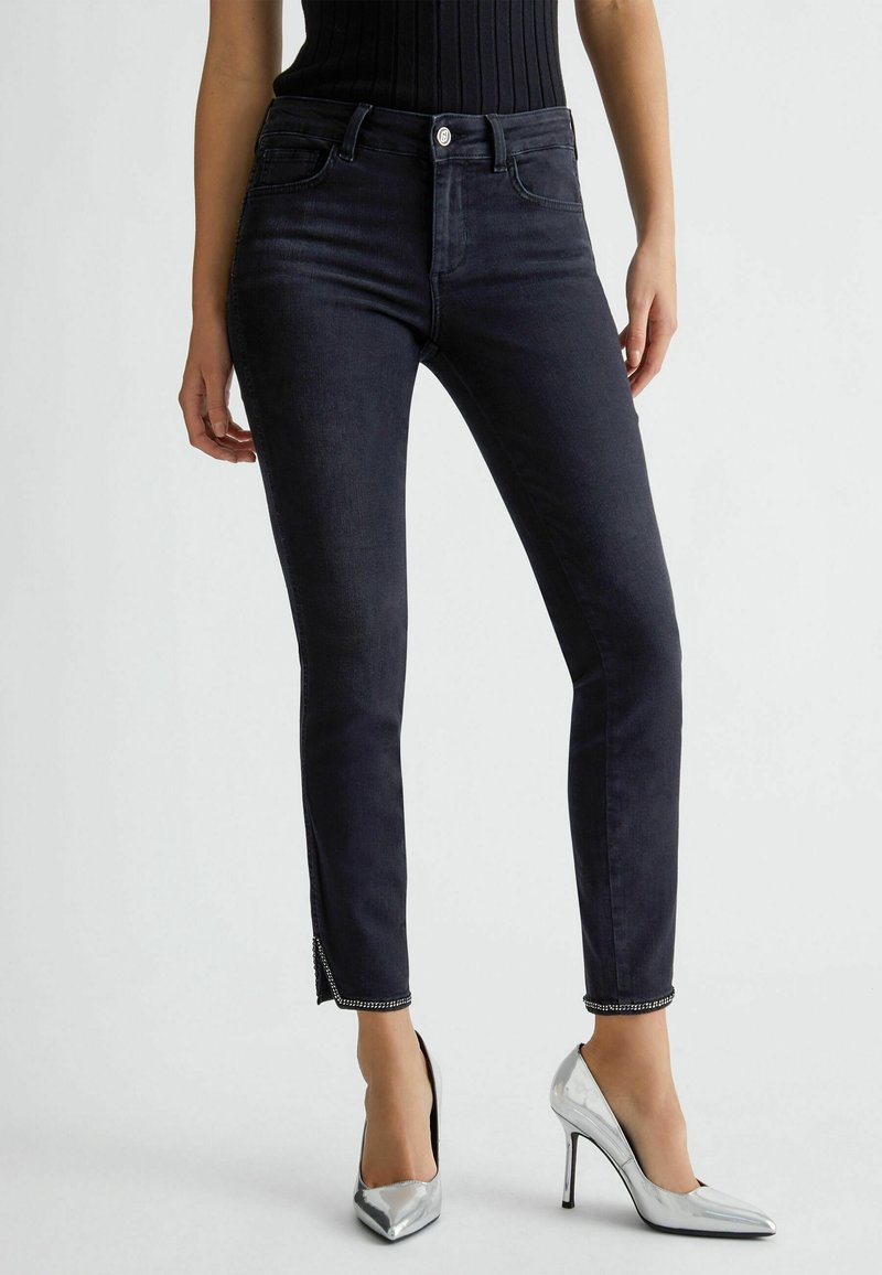 Jeans ajustados de mezclilla negro de cintura alta, con aberturas laterales en los tobillos y detalles de herrajes plateados. Combinados con zapatos de tacón alto metálicos.