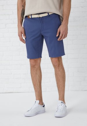 Mann trägt blaue knielange Shorts, weißen geflochtenen Gürtel, beigefarbenes Oberteil und weiße Sneaker, steht vor einer weißen Ziegelsteinmauer.