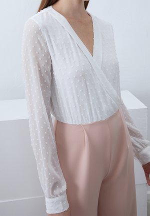 Witte blouse met gestructureerde polkadots, diepe V-hals en lange doorschijnende mouwen, gecombineerd met een lichtroze broek met hoge taille.