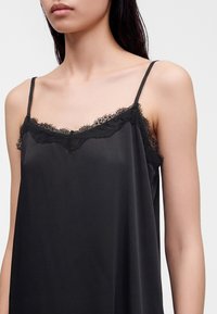 Jeune femme portant un caraco en satin noir avec fines bretelles et bordure en dentelle le long de l'encolure et des emmanchures.