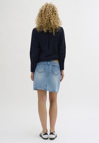Denim skirt features a frayed hem and two back pockets. Paired with a dark blouse and black-and-white sneakers, shot from the back.