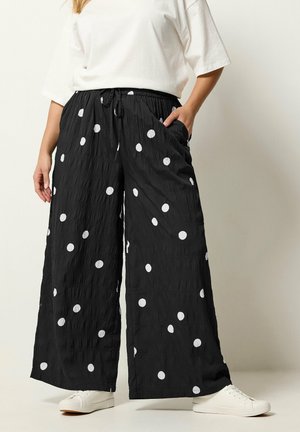 Pantalon large noir à pois blancs, taille élastique avec cordon, associé à des baskets blanches et un haut blanc à manches courtes.