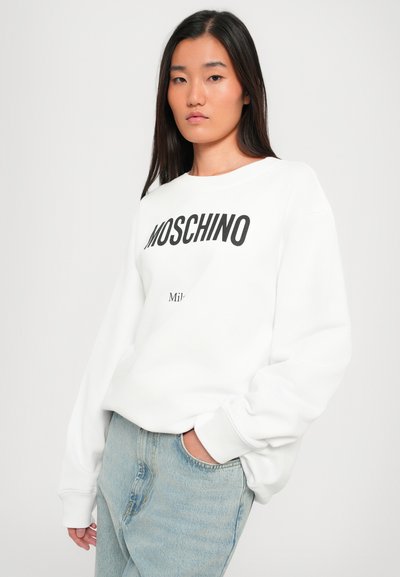 Vit sweatshirt med svart "MOSCHINO"-logotyp och mindre text nedanför. Har rund halsringning, långa ärmar och mjukt tyg. Matchas med ljusa jeans.