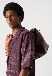 Lila Kurzarmshirt mit einem Kartenprint in Beige. Eine braune Lederhandtasche hängt über der Schulter und hat einen Kordelzugverschluss.