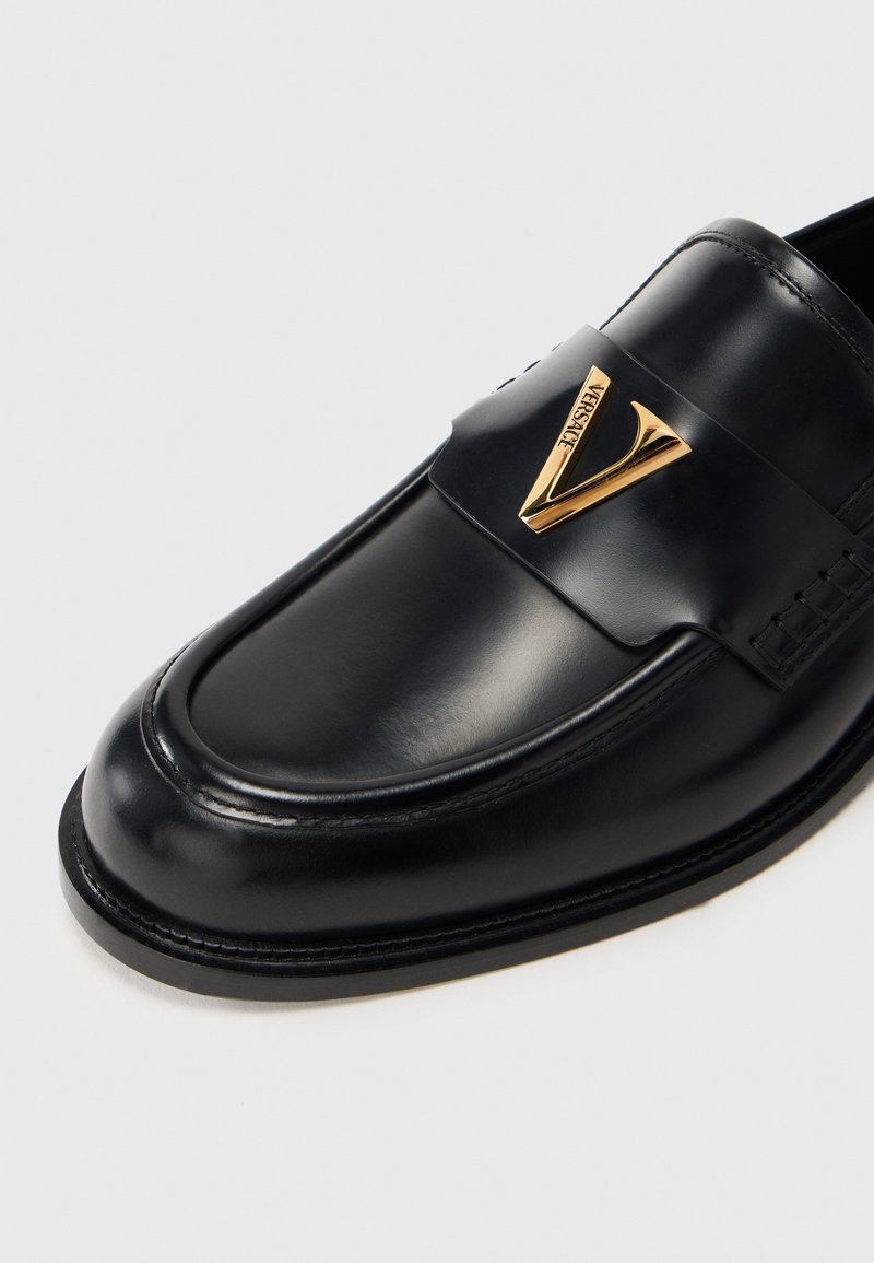 Mocassin en cuir noir avec un accent logo doré, bout arrondi, détails cousus sur les côtés et une finition lisse et brillante.