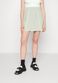 Světlounce zelená mini sukně s vlnitým lemem a bílým lemem; spárována s černými mokasíny s bílým akcentem a detailem třásní.