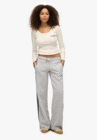 Vrouw met krullend haar, gekleed in een cropped crèmekleurige top met lange mouwen en grijze wijde sweatpants met zijstreep en beige sneakers, staand met de handen in de zakken.