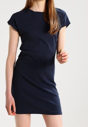 Kobieta w granatowej, dopasowanej sukience z krótkim rękawem stojąca na jednolitym, jasnym tle.