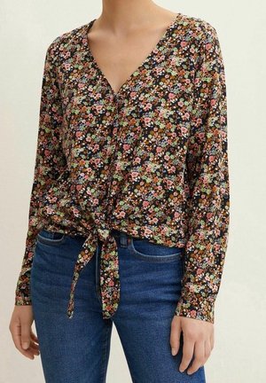 Bluzka z długim rękawem w kwiaty w kolorze różowym, zielonym i białym na czarnym tle, zawiązana z przodu na supeł, noszona z niebieskimi dżinsami z denimu.