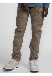 Beige cargobroek met meerdere zijzakken, een relaxte pasvorm en subtiele kreukels, gecombineerd met grijze en witte sneakers met tan accenten.
