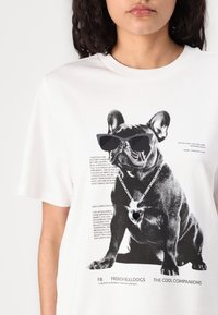 White t-shirt with a black and white graphic of a French Bulldog wearing sunglasses and a heart necklace, with text detailing the breed.