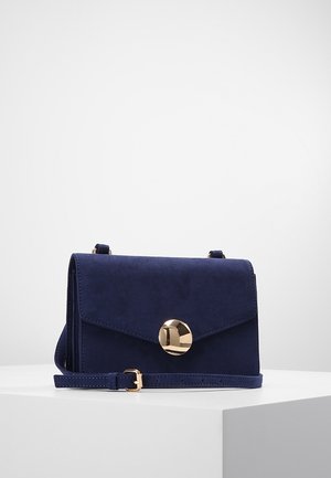 Sac bandoulière en daim bleu marine avec une fermeture ronde en or et une sangle réglable, présenté sur un socle blanc sur un fond uni.