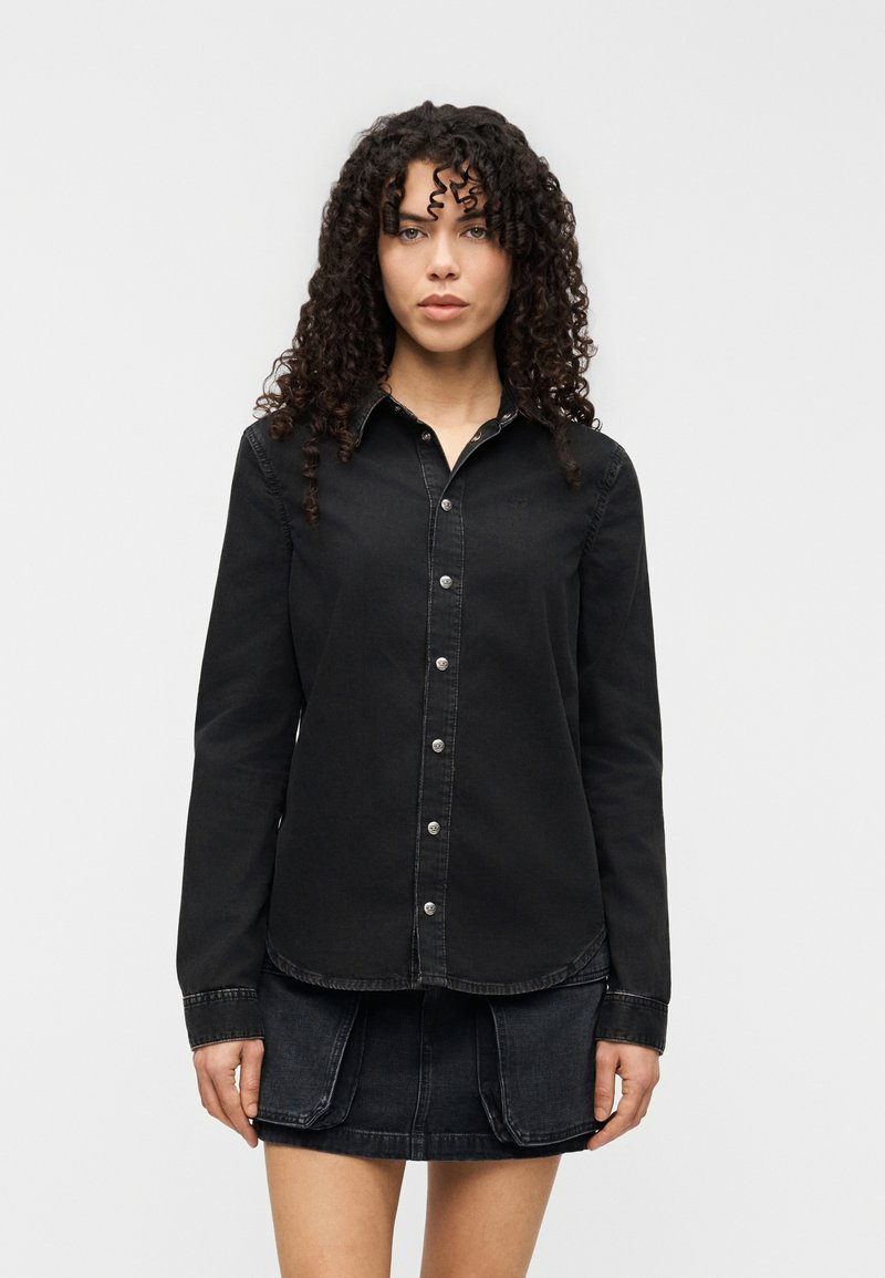 Μαύρο τζιν πουκάμισο με κλείσιμο με κουμπιά και μακρυά μανίκια, φορεμένο με φούστα από το ίδιο ύφασμα τζιν. Το ύφασμα έχει ομαλή υφή και διακριτικές ραφές.