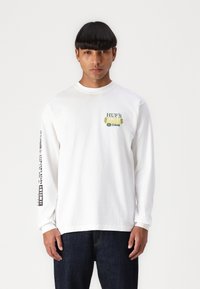 Hvid langærmet t-shirt med et grønt grafisk tryk på brystet og sort tekst langs den venstre ærme, lavet af bomuldsstof.