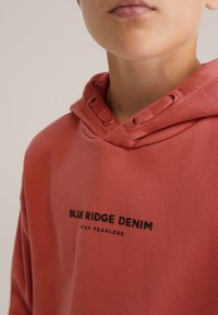 Sudadera ligera roja hecha de algodón, con un bolsillo tipo canguro, capucha con cordón y texto negro que dice "BLUE RIDGE DENIM STAY FEARLESS".