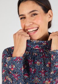 Blusa con motivo floreale su base blu navy, colletto alto e maniche lunghe. Presenta intricati disegni floreali in rosso, beige e bianco. Tessuto dalla texture morbida.