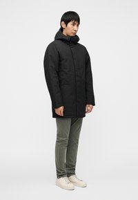 Μαύρο επενδυμένο μπουφάν με ψηλή γιακά και κουκούλα, με μπροστινό φερμουάρ, πλαϊνές τσέπες, φορεμένο με παντελόνι χακί και ανοιχτόχρωμα αθλητικά παπούτσια.