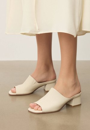 Pėdos dėvinčios kremiškos spalvos atvirų pirštų aukštakulnius su tvirtu kulnu, derinamus su šviesiai kremine midi ilgio suknele, stovinčios ant lygaus grindinio.