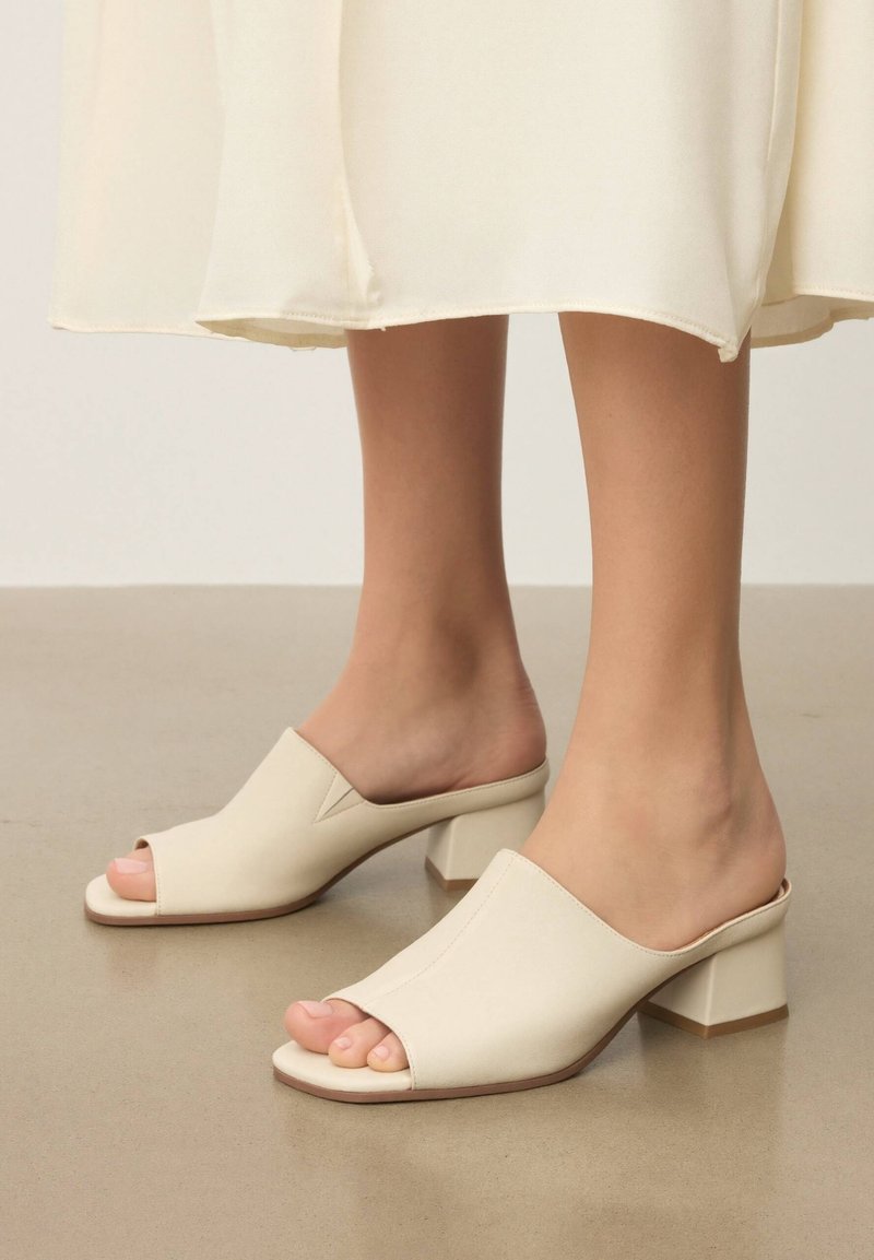 Pieds portant des mules à talons ouverts de couleur crème avec un talon carré, associées à une jupe midi crème clair, debout sur un sol lisse.