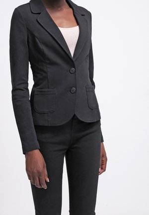 Vrouw draagt een nauwsluitend zwart colbert met twee knopen en voorzakken, over een wit topje en zwarte broek, staand tegen een effen achtergrond.