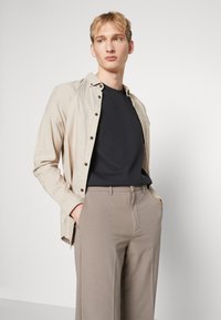 Camicia button-up beige chiaro sopra una maglietta nera a girocollo, abbinata a pantaloni grigi chiari su misura, tutti caratterizzati da texture lisce.