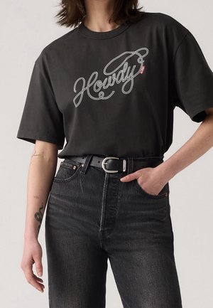 Henkilö pukeutunut mustaan t-paitaan, jossa "Howdy"-teksti köyden tyylisellä fontilla, laitetttu korkeavyötäröisten mustien farkkujen sisään mustalla vyöllä ja hopeisella soljella.