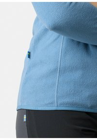 Jasny niebieski polarowy kurtka o miękkiej teksturze, z małym akcentem w postaci szwedzkiej flagi i etykietą tekstową wzdłuż dołu.