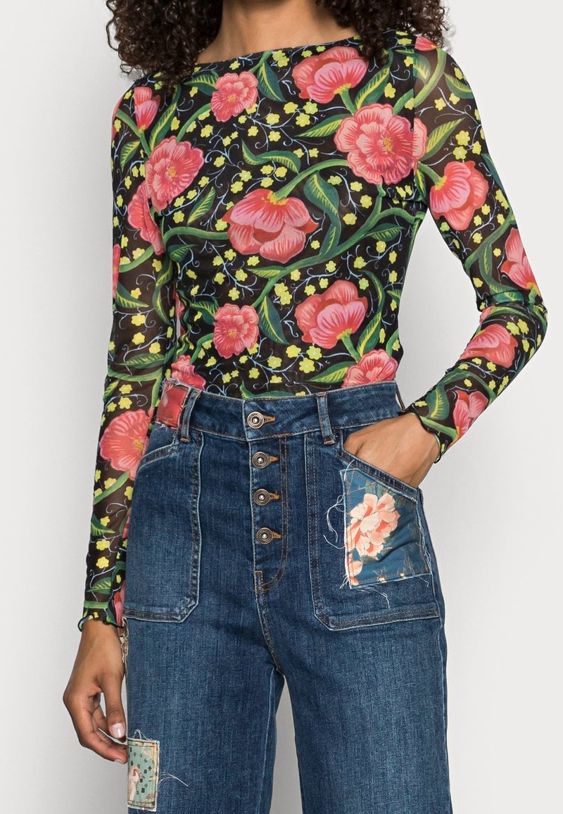 Femme portant un haut noir transparent à manches longues avec un grand imprimé floral rose et un jean bleu taille haute avec des poches patch florales.