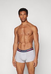 Graue Baumwoll-Boxershorts mit einem marineblauen Bund, der rot-weiße Streifen aufweist, mit dem Aufdruck "LACOSTE", enganliegendes Design mit glatter Textur.