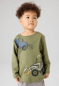 Jaunas vaikas su garbanotais plaukais, dėvintis alyvuogių žalios spalvos ilgomis rankovėmis marškinėlį su mėlynais ir baltais statybinių mašinų atspaudais.