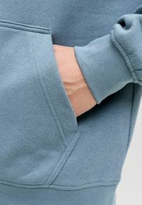 Felpa con cappuccio azzurro chiaro con tasca a marsupio, bordi cuciti e polsini a costine. Texture liscia con design a vestibilità rilassata.