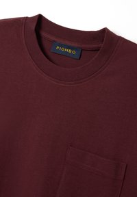 Camiseta burdeos de algodón, con un cuello redondo y un bolsillo en el pecho. La etiqueta muestra "PIOMBO" en texto amarillo sobre un fondo oscuro.