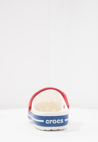 Crocs vermelhos e azuis com uma alça traseira, apresentando um interior branco e sola texturizada, com listras horizontais e o logotipo da marca na lateral.