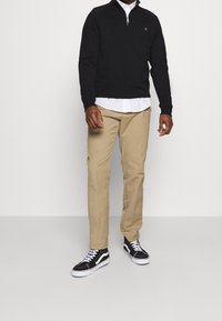 Sudadera de cremallera negra, camisa blanca de cuello debajo, pantalones caqui, zapatillas en blanco y negro con cordones. Las características son mínimas y casuales.