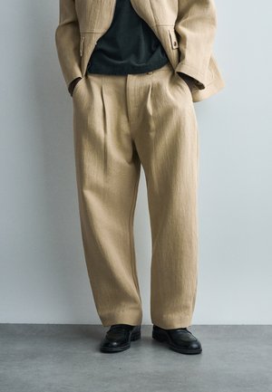 Άνδρας με μπεζ πλισέ παντελόνι και ταιριαστό σακάκι, μαύρα μοκασίνια και μαύρο πουκάμισο, με τα χέρια στις τσέπες, στέκεται σε γκρίζο δάπεδο.