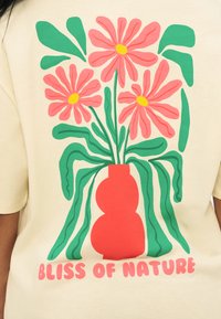 Τρία μεγάλα ροζ λουλούδια με κίτρινα κέντρα και πράσινα φύλλα σε κόκκινο βάζο, με κείμενο "BLISS OF NATURE" από κάτω σε μπεζ ύφασμα.