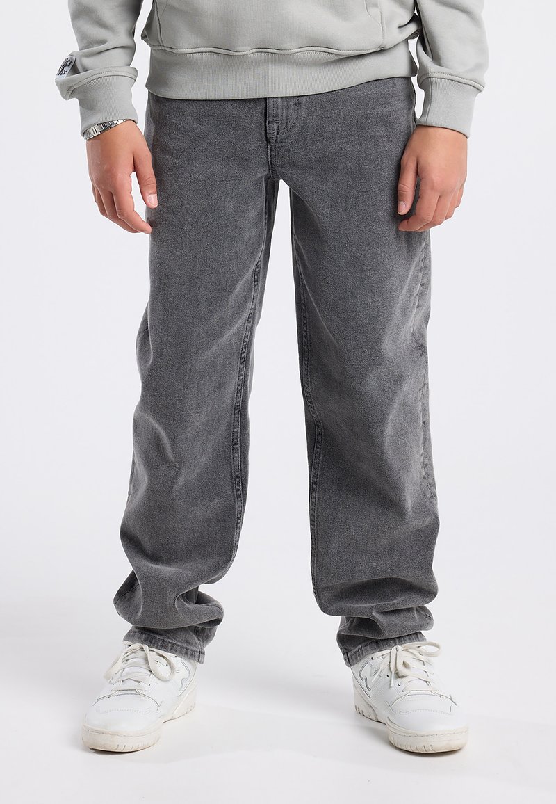 Pantaloni in denim grigio con vestibilità rilassata, caratterizzati da una superficie texturizzata. Abbinati a sneakers bianche, con lacci e un aspetto leggermente usurato.