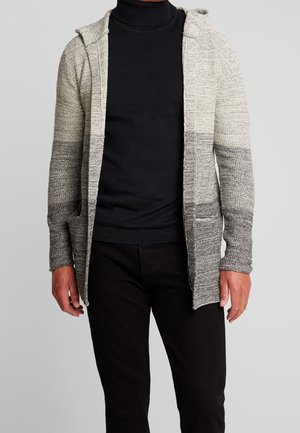 Homme portant un cardigan à capuche en tricot gris ombré par-dessus un col roulé noir et un pantalon noir, debout devant un fond uni.